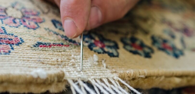 Untitled-3-1-ezgifcom-jpg-to-webp-converter-e1767449494237 - Gohari Oriental Rugs Service Close-up of a hand using a needle to repair a frayed edge of a beige patterned rug, stitching the fringe closed.