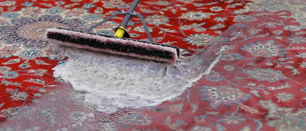 Untitled-eee1-ezgifcom-jpg-to-webp-converter - Gohari Oriental Rugs Service A carpet cleaning tool with a pink roller scrubs a red floral rug, spraying white foam as it cleans.