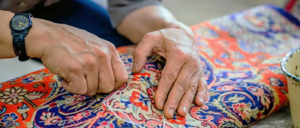 persian-rug-repair-ezgifcom-jpg-to-webp-converter-e1767449434314 - Gohari Oriental Rugs Service Close-up of a person’s hands mending a colorful patterned fabric with a needle and thread, a black wristwatch visible on the wrist.